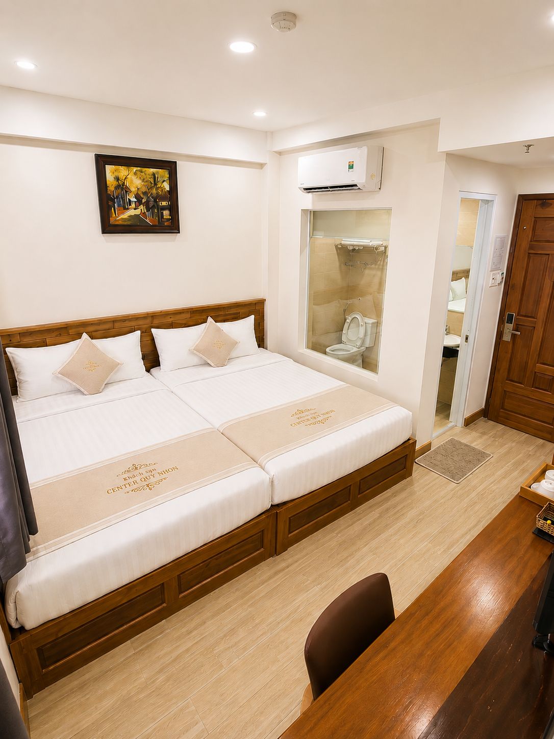 A hotel room with a double bed, a wooden desk, a chair, a wardrobe, and a bathroom visible through a glass door.