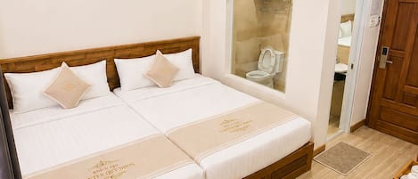 A hotel room with a double bed, a wooden desk, a chair, a wardrobe, and a bathroom visible through a glass door.
