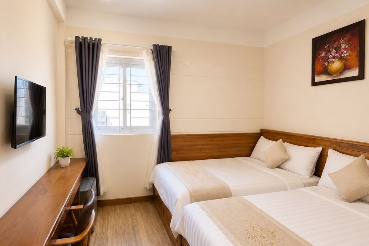 A hotel room with a wooden desk, two beds, a television, and a window with curtains.