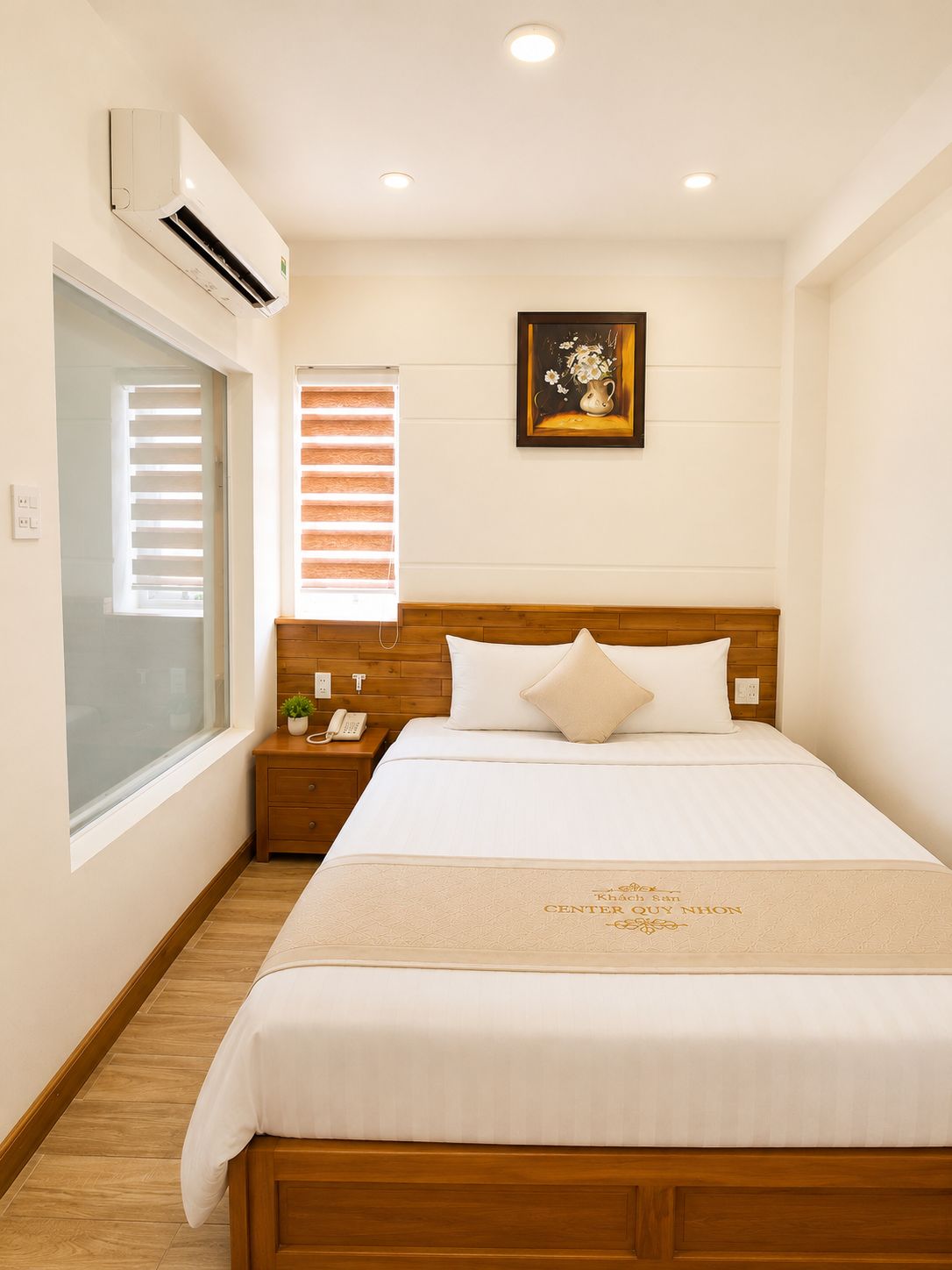 A hotel room with a bed, bedside table, nightstand, wall-mounted air conditioner, window with blinds, and a framed picture on the wall.