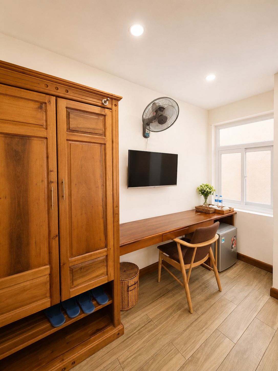 Quarto com armário de madeira, escrivaninha, cadeira, televisão, ventilador, mini-geladeira e janela.
