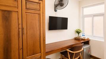 Quarto com armário de madeira, escrivaninha, cadeira, televisão, ventilador, mini-geladeira e janela.