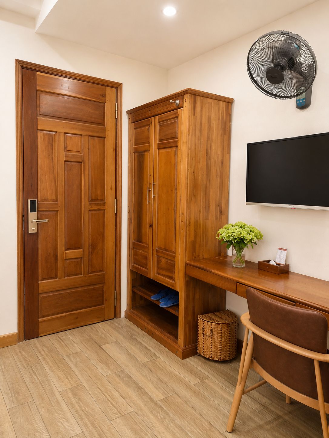 Quarto com guarda-roupa de madeira, escrivaninha com cadeira, televisão e ventoinha fixada na parede.