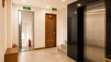 Un hall de hotel con una puerta de madera que indica 'EXIT', un extintor, una escalera y un ascensor.