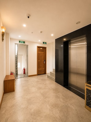 A hotel lobby with a wooden door marked 'EXIT,' a fire extinguisher, a staircase, and an elevator.