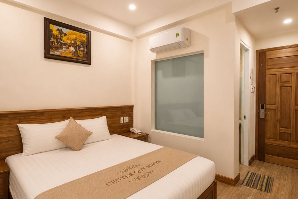 A hotel room with a bed, a painting on the wall, an air conditioning unit, and a wooden door.