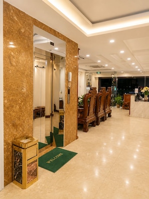 A hotel lobby with a reception desk, a green welcome mat, and a row of wooden chairs.