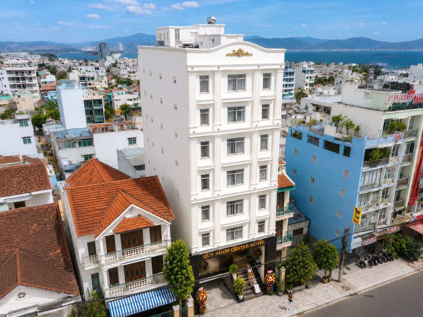 Hotel de vários andares com entrada central, rodeado por outros edifícios, e vista para o mar e montanhas ao fundo.