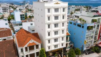 Hotel de vários andares com entrada central, rodeado por outros edifícios, e vista para o mar e montanhas ao fundo.