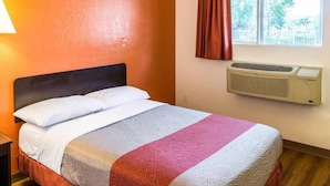 Un lit simple avec une couverture rayée blanche et rose, un climatiseur fixé au mur et une fenêtre avec un rideau.