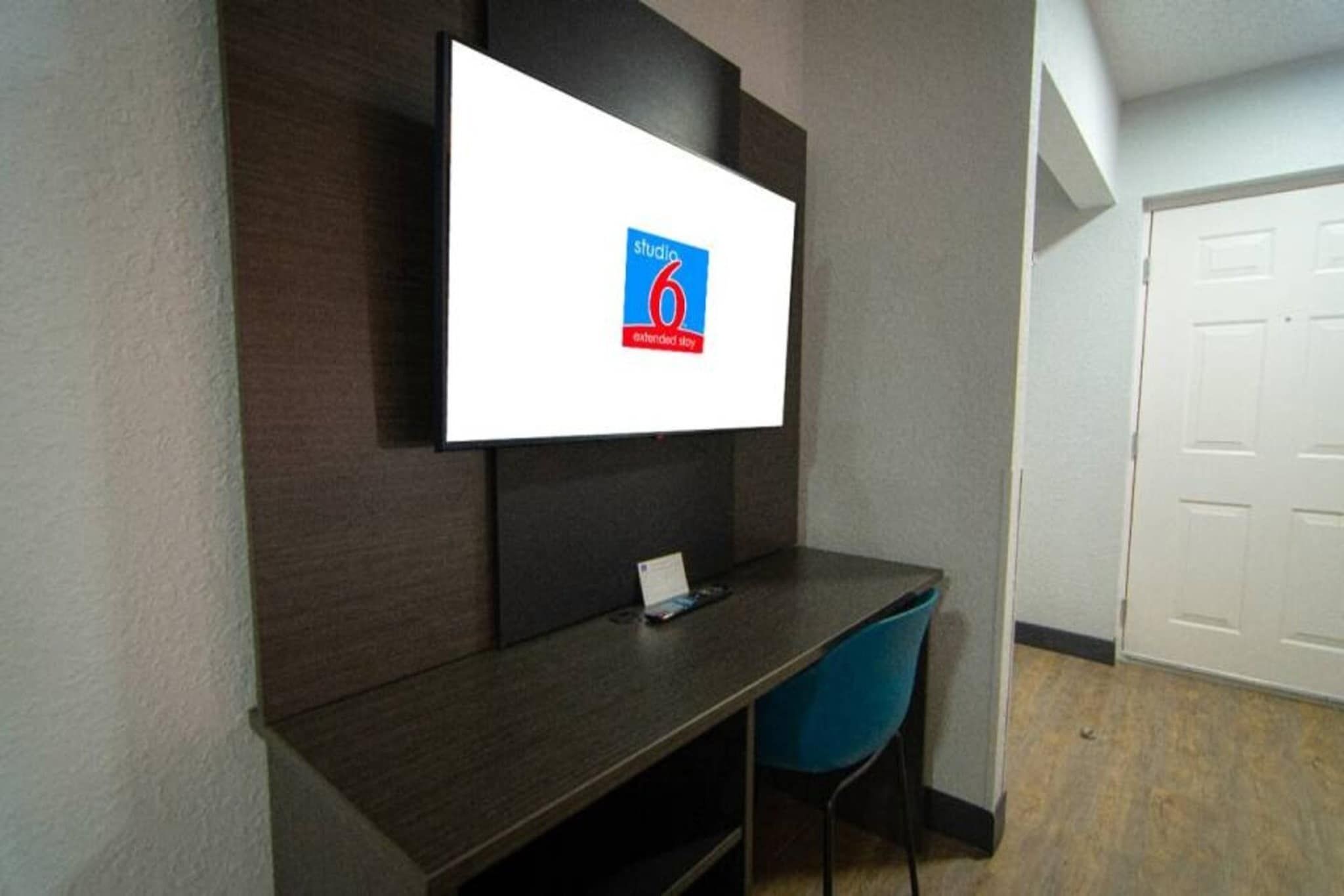 A room with a desk, a chair, a flat-screen TV mounted on the wall, and a closed door.