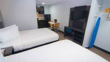 A hotel room with two beds, a desk, a chair, a TV, and a kitchenette in the background.