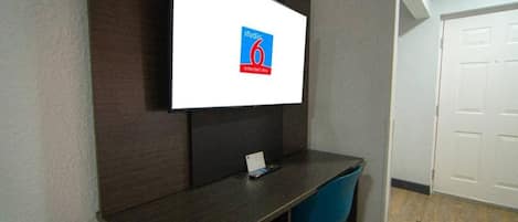A room with a desk, a chair, a flat-screen TV mounted on the wall, and a closed door.