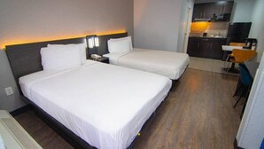 Habitación de hotel con dos camas individuales, un escritorio con silla y una kitchenette al fondo.