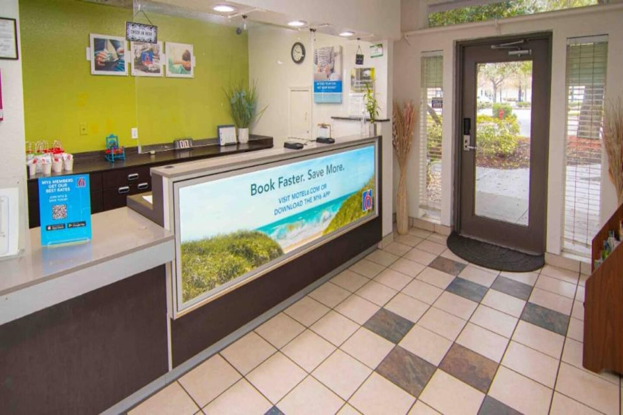 Zona de recepción con una pantalla digital mostrando un mensaje de reserva, un mostrador con un cartel para miembros y un suelo de baldosas.