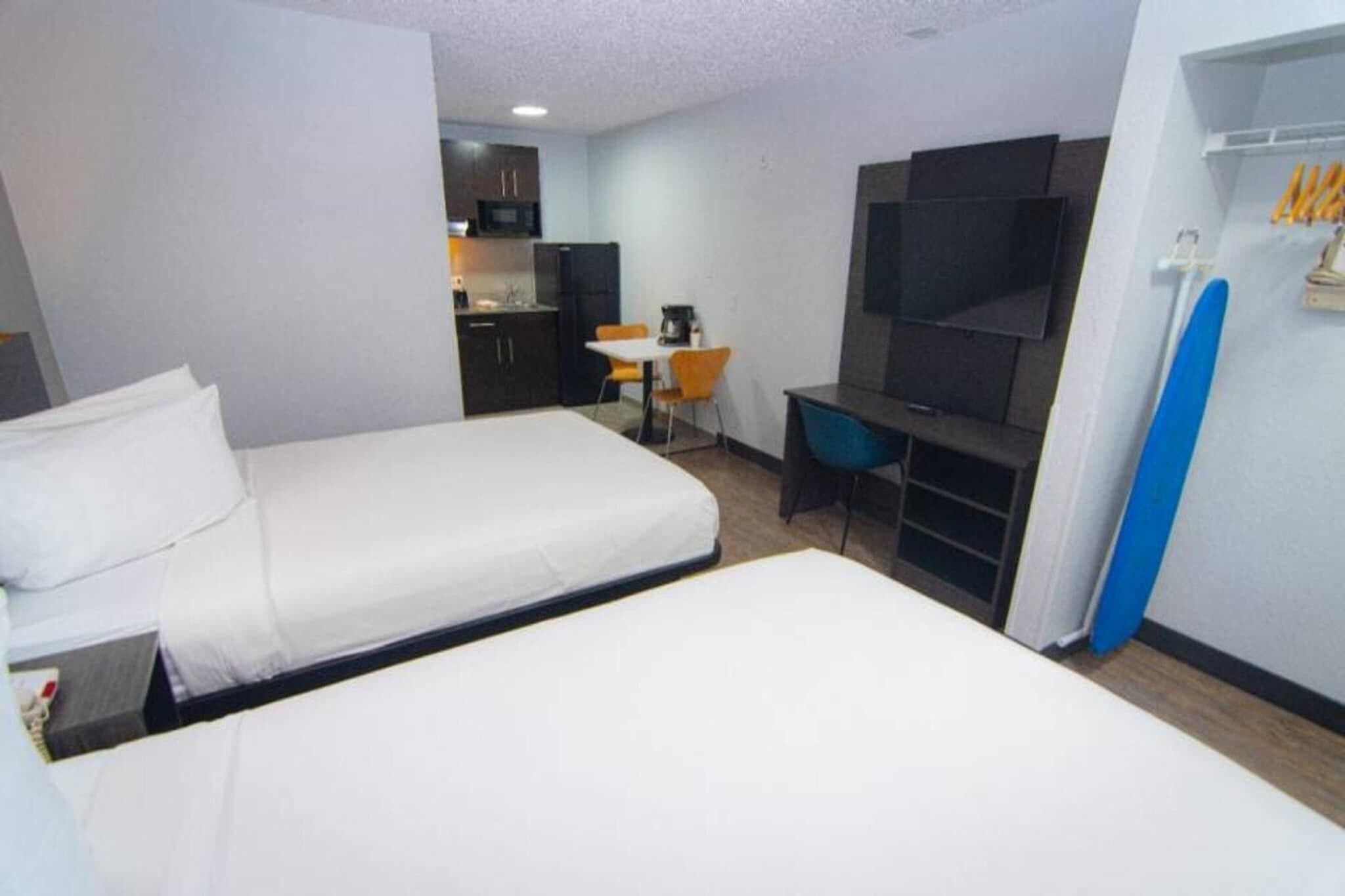 A hotel room with two beds, a desk, a chair, a TV, and a kitchenette in the background.