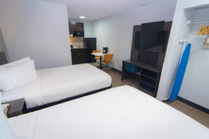 A hotel room with two beds, a desk, a chair, a TV, and a kitchenette in the background.