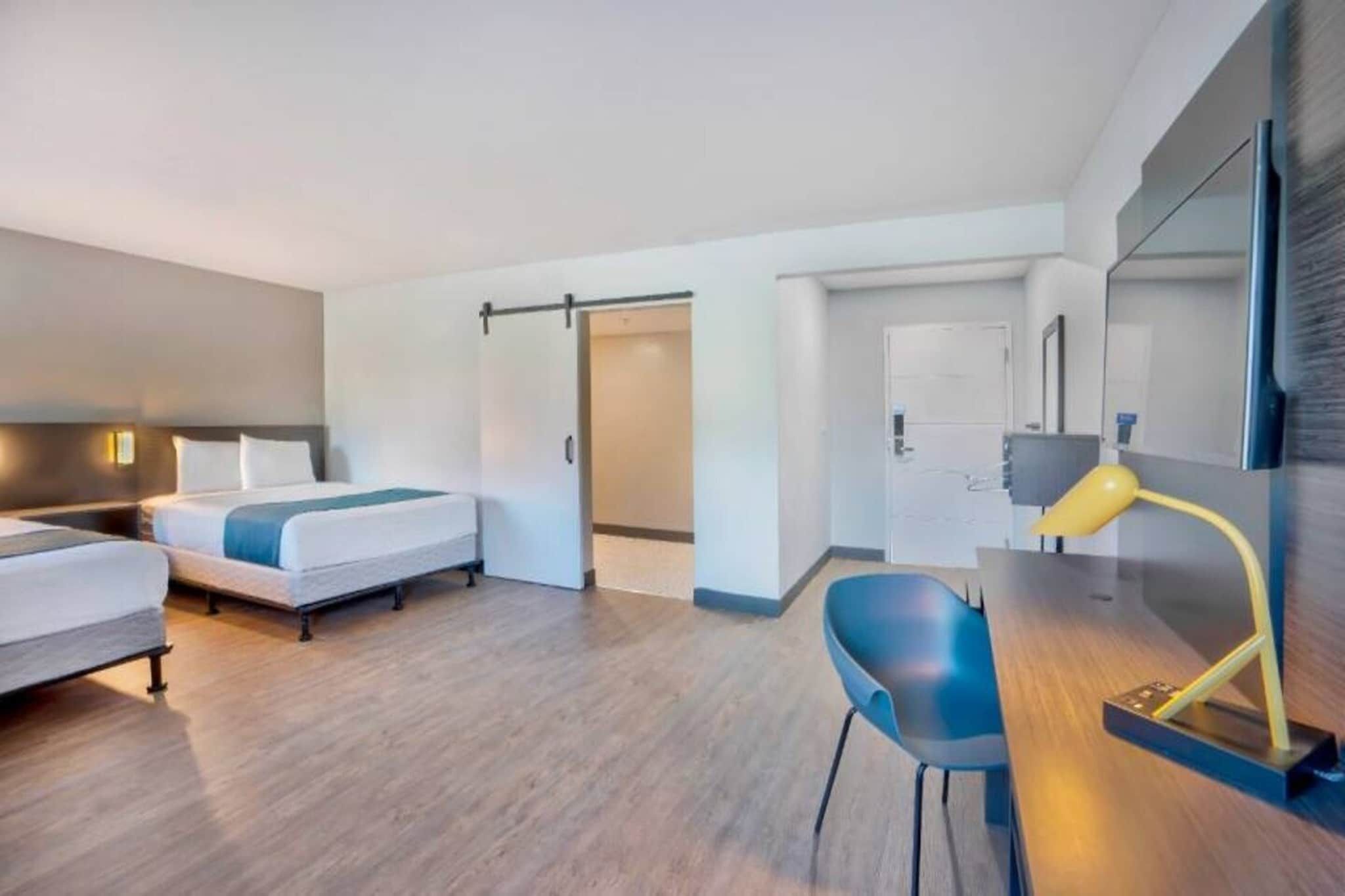 A hotel room with two beds, a desk with a chair, a mirror, and a door leading to a bathroom.