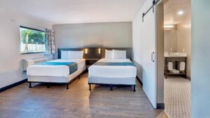 Una camera d'albergo con due letti, una grande finestra, un bagno con lavandino e specchio, e una porta che dà su un'altra stanza.