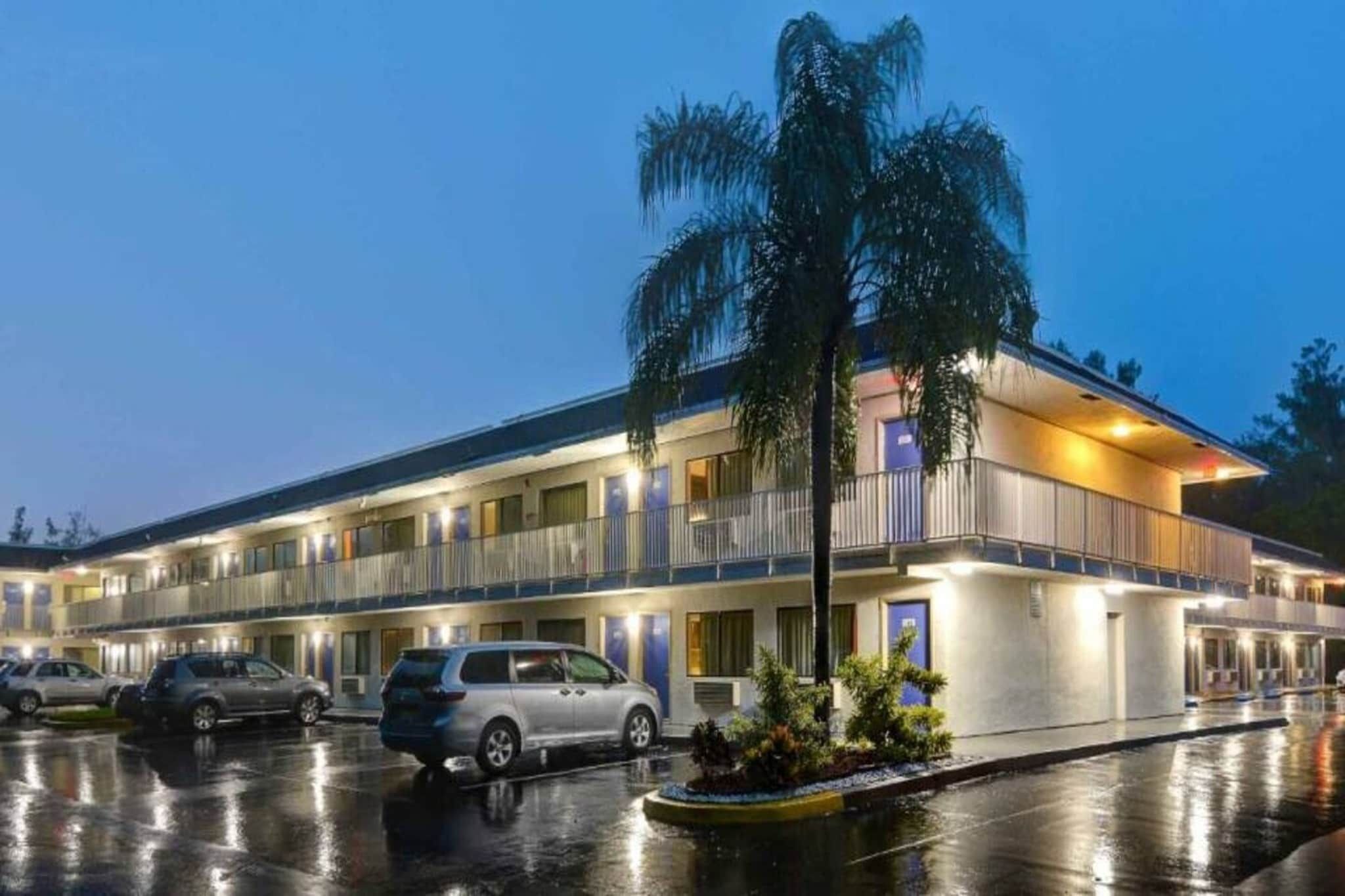 Un motel de varios pisos con estacionamiento y una palmera en primer plano.