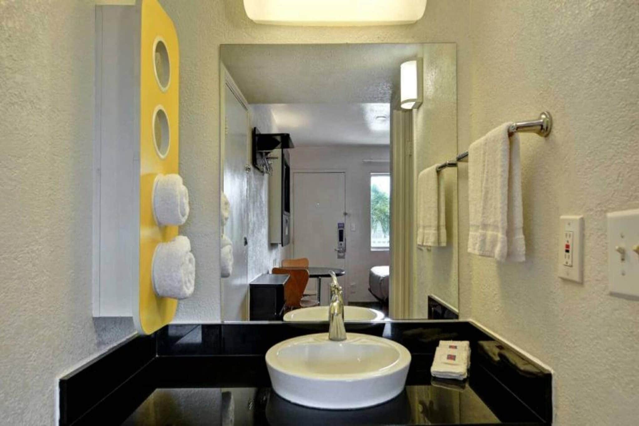 Un baño de hotel con encimera negra, lavamanos blanco, espejo y toallero con toallas.
