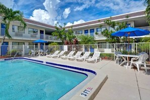 A swimming pool with lounge chairs and umbrellas, surrounded by a fenced area with a building in the background.
