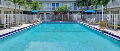 A swimming pool with a white fence, surrounded by palm trees and a building with balconies.