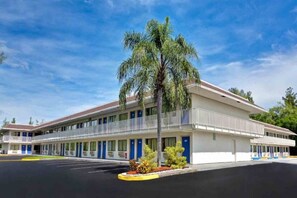 Un motel composé d’un bâtiment à deux étages, d’un espace de stationnement couvert et d’un palmier à l’avant.