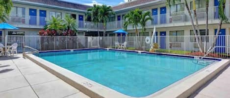 A rectangular swimming pool with a tiled edge, surrounded by a white fence, and adjacent to a building with balconies and windows.