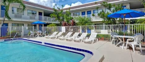 A swimming pool with lounge chairs and umbrellas, surrounded by a fenced area with a building in the background.