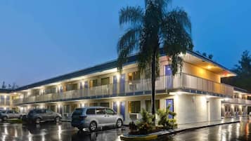 Un motel de varios pisos con estacionamiento, coches aparcados y una palmera en primer plano.