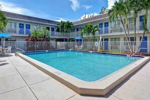 Una piscina rectangular con un bordeado de baldosas, una zona vallada y un edificio con balcones al fondo.