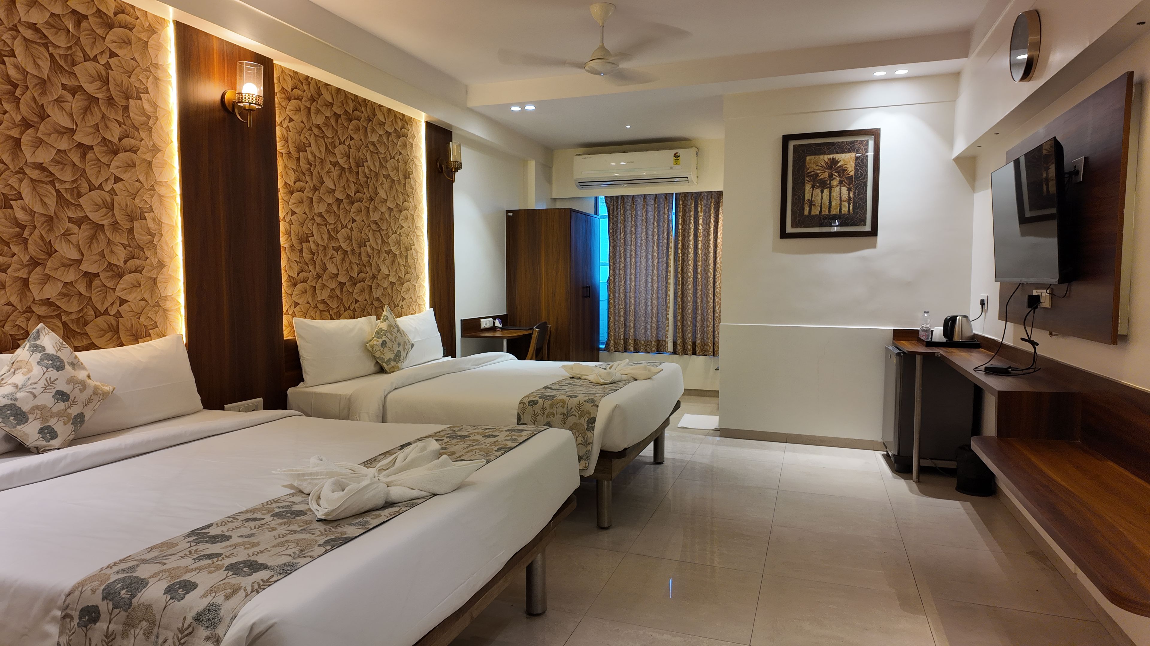 A hotel room with two beds, a desk, a TV, and an air conditioning unit.