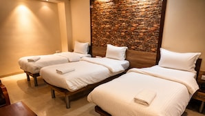 Ein Hotelzimmer mit drei Einzelbetten, jedes mit einem weißen Kissen und einem gefalteten Handtuch. Das Zimmer verfügt über eine Steinwand und ein hölzernes Kopfteil.