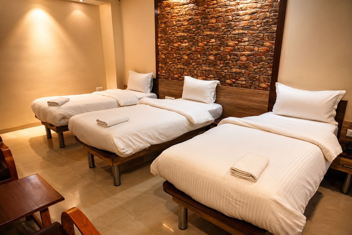 A hotel room with three single beds, each with a white pillow and a folded towel. The room has a stone wall feature and a wooden headboard.