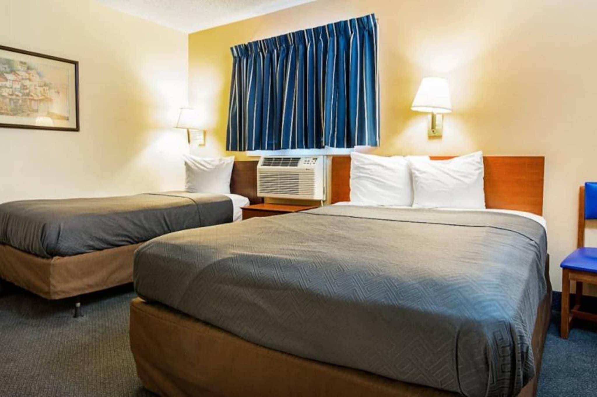 A hotel room with two beds, a window with curtains, an air conditioning unit, and a lamp.
