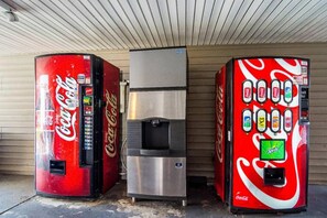 Três máquinas de venda automática de bebidas, uma com topo prateado e duas com marcação vermelha e branca, diante de uma parede lisa.