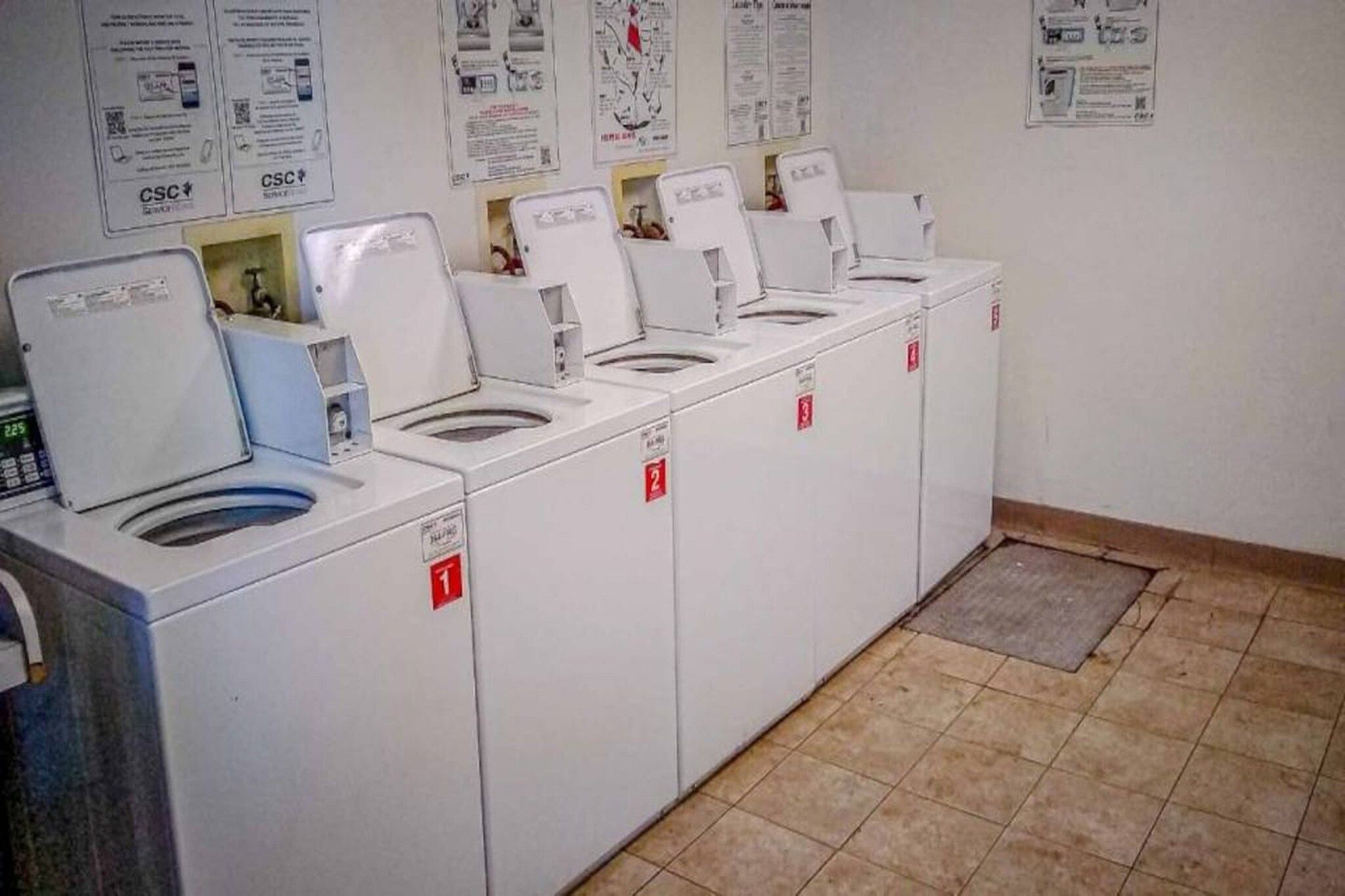 Eine Reihe von weißen Wäschemaschinen mit nummerierten Türen, ein gefliester Boden und Informationsplakate an der Wand.
