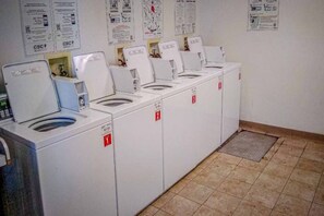 Fileira de máquinas de lavar roupa brancas, com portas numeradas, chão em mosaico e cartazes informativos na parede.