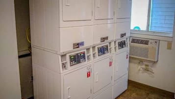 A laundry room with stacked washing machines and dryers, a window with blinds, and a visible electrical outlet.
