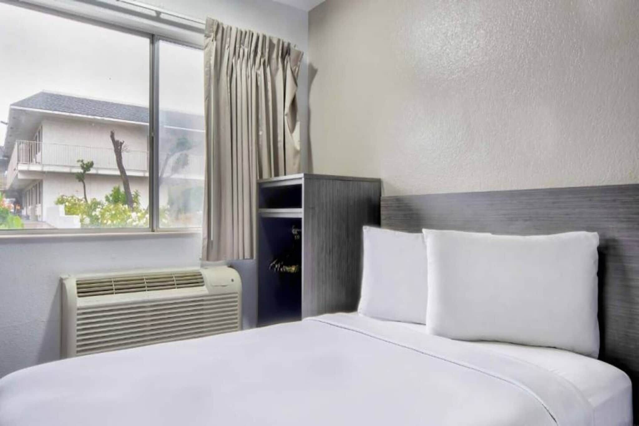 A hotel room with a bed, a window with curtains, a wall-mounted heater, and a view of the outside.