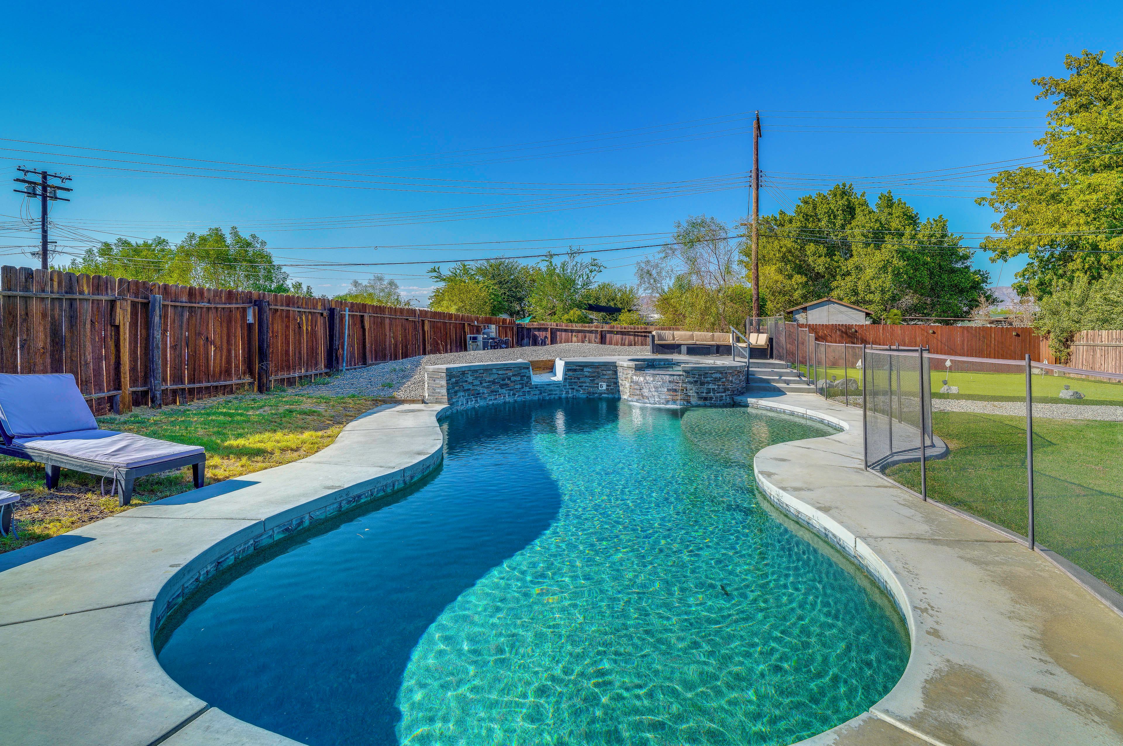 A clear blue swimming pool with a curved design, surrounded by a wooden fence, and a small shed in the background.