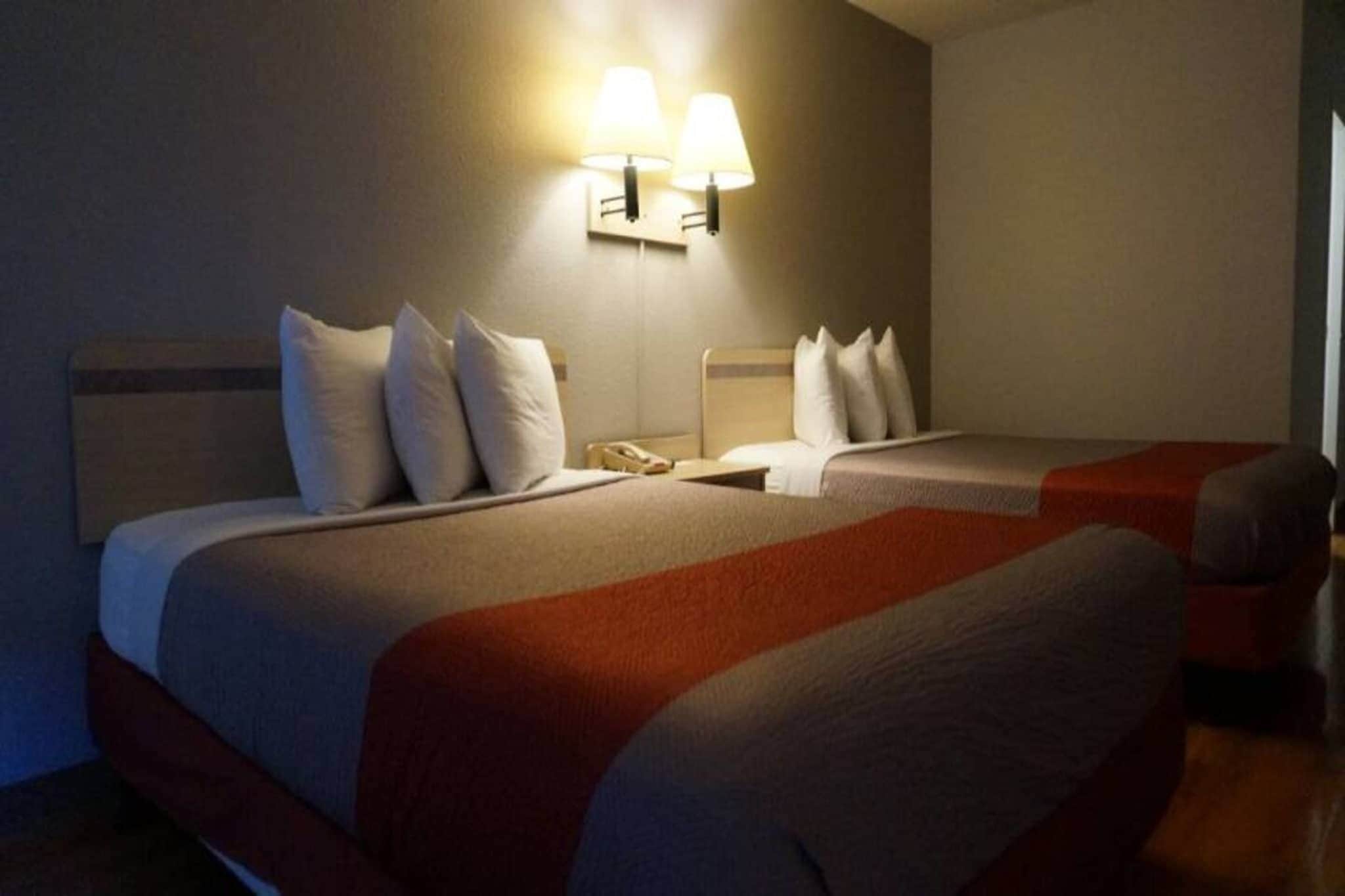 Hotellihuoneessa on kaksi sänkyä, joissa on valkoiset vuodevaatteet ja punaiset koriste-elementit. Huoneessa on kaksi seinään kiinnitettyä lamppua, jotka valaisevat tilaa.