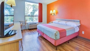 Habitación de hotel con cama, escritorio con lámpara, silla, ventana y unidad de aire acondicionado.