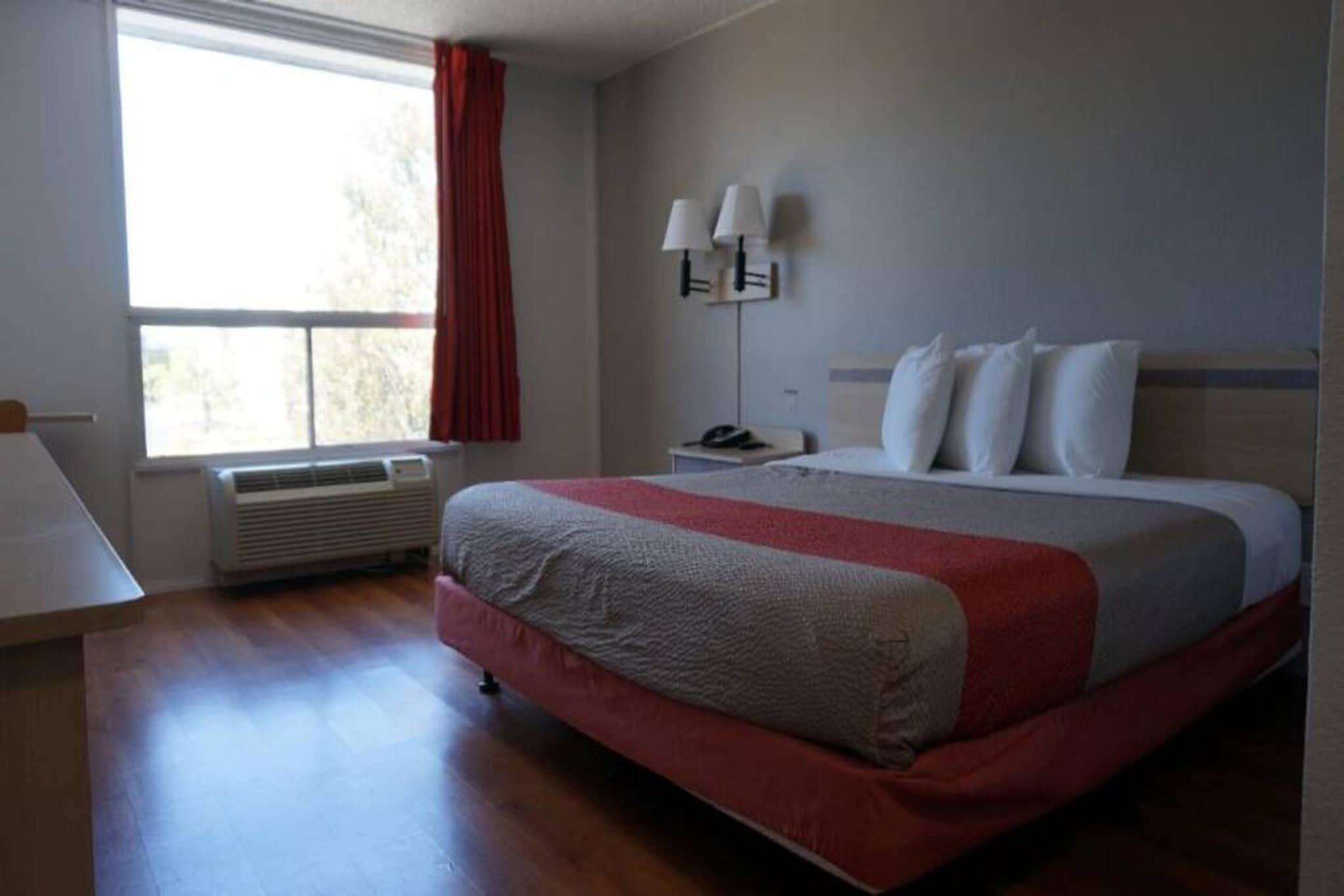 A hotel room with a large bed, a desk, a chair, a window with curtains, and a wall-mounted lamp.