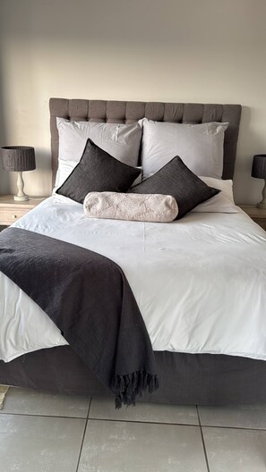 Una cama con cabecera tapizada en gris, ropa de cama blanca y una manta gris oscura.