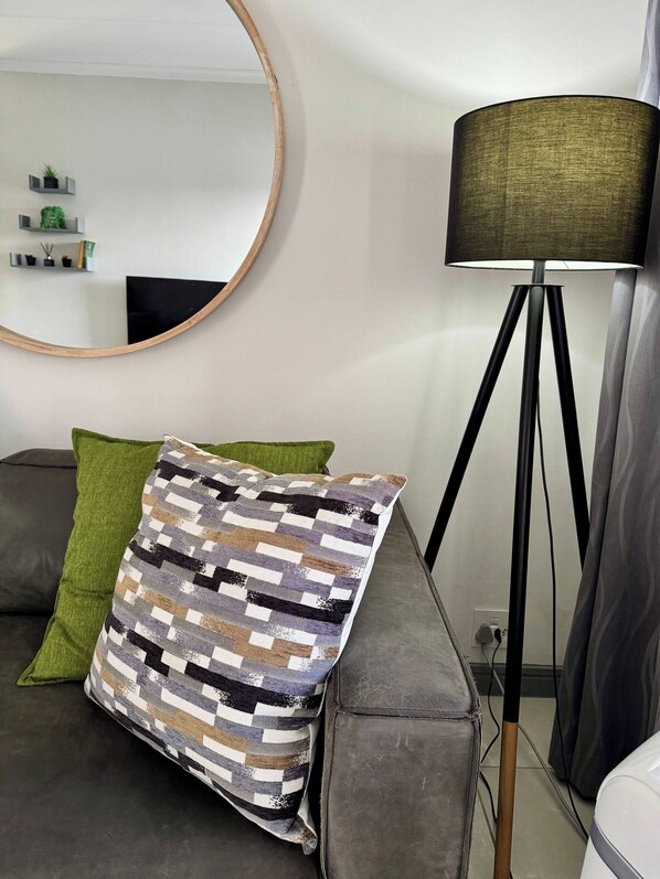 Una sala de estar moderna con un sofá gris, un cojín estampado, un cojín verde, un espejo redondo y una lámpara de pie.