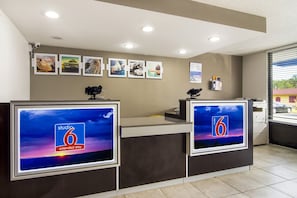 Mostrador de recepción de hotel con dos monitores mostrando el logotipo de 'Studio 6 Extended Stay', un tablón de anuncios con varias fotos y una ventana con persianas.