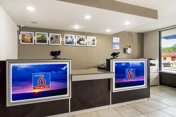 A hotel reception desk with two monitors displaying the 'Studio 6 Extended Stay' logo, a bulletin board with various photos, and a window with blinds.
