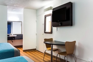 Une chambre d’hôtel avec un lit, un bureau et une chaise, un téléviseur fixé au mur, et une salle de bain visible à travers une porte ouverte.